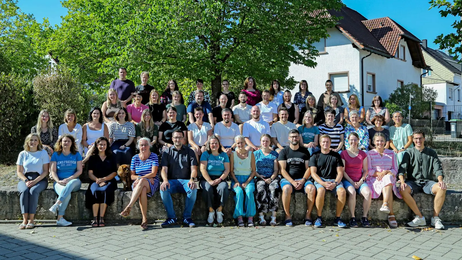 Gruppenfoto des Kollegiums auf einer Steinmauer und Treppe vor Bäumen und einem Wohnhaus bei Sonnenschein