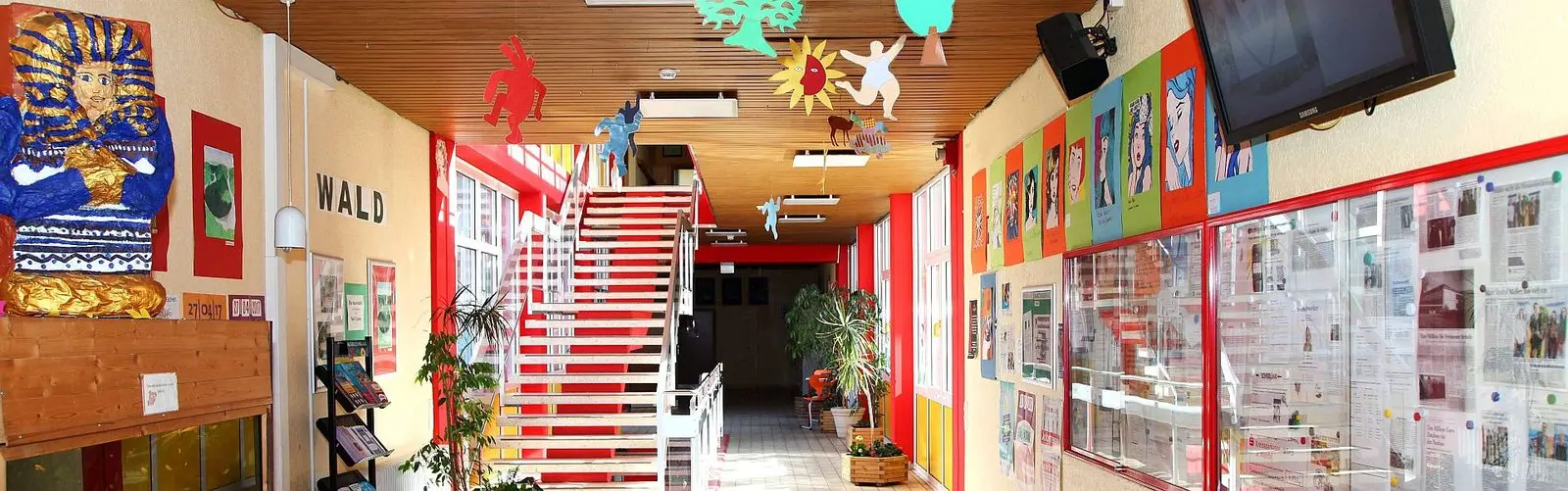 Schulflur mit roter Treppe, bunten Bildern an den Wänden und Schaukästen