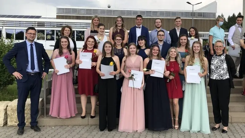 Abiturientinnen und Abiturienten mit Zeugnismappen und Rosen auf einer Treppe vor dem Schulgebäude