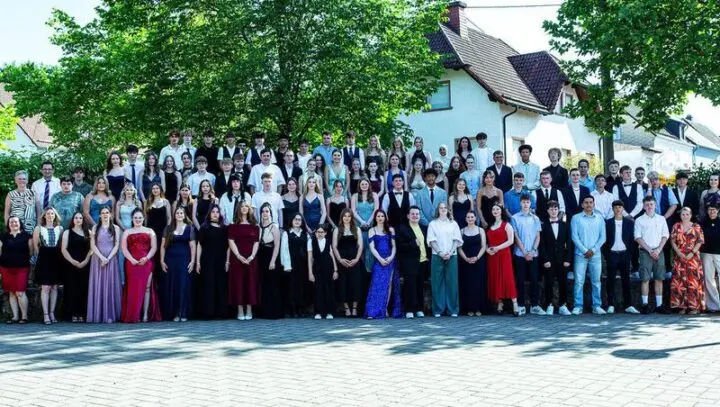 Großes Gruppenfoto der Absolventinnen und Absolventen in festlicher Kleidung vor Bäumen und Häusern