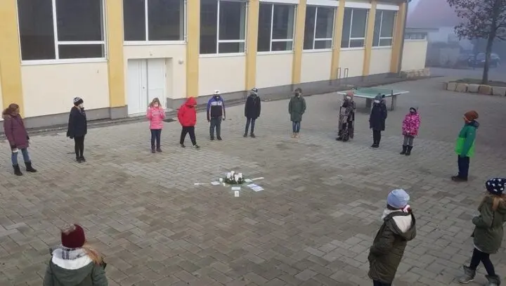 Kinder mit Masken stehen mit Abstand im Kreis um einen Adventskranz auf dem Schulhof