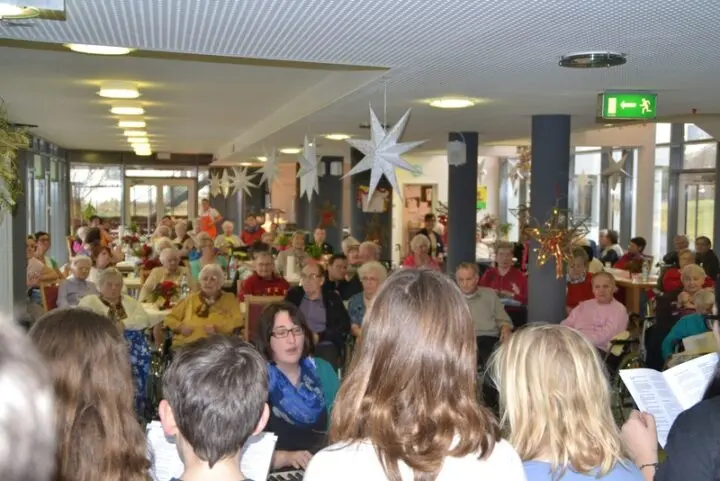 Chor mit Notenblättern singt vor Seniorinnen und Senioren in einem weihnachtlich geschmückten Saal