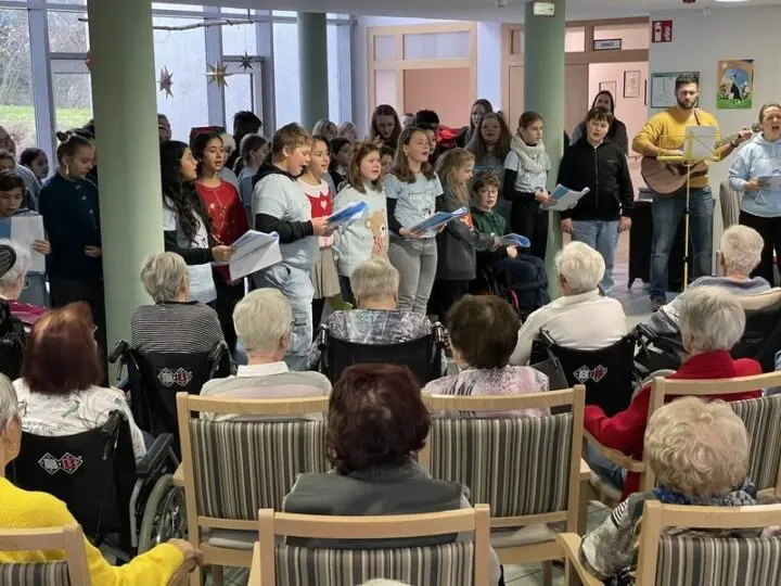 Schülerinnen und Schüler singen mit Liedblättern vor Seniorinnen und Senioren, ein Mann spielt Gitarre