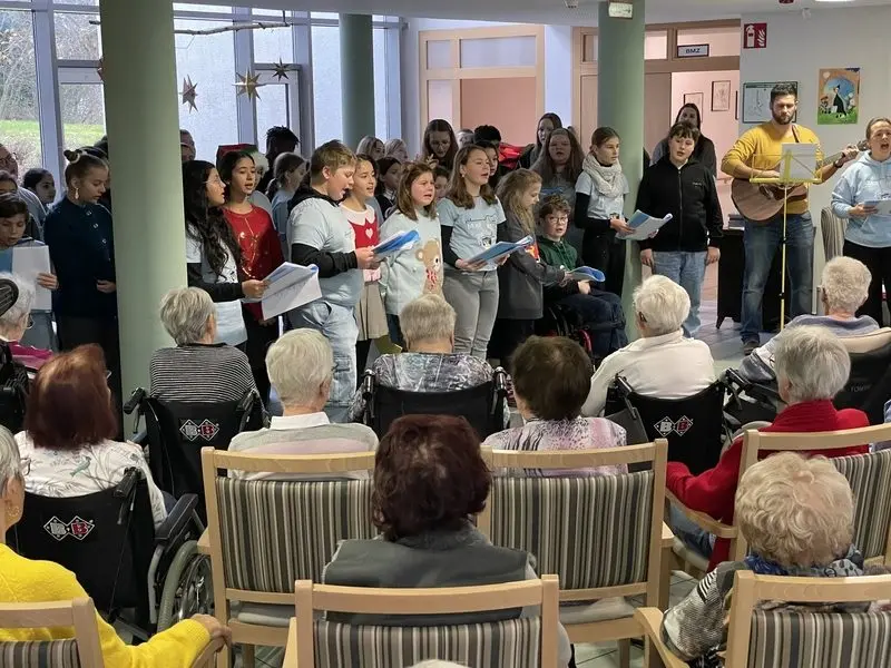 Schülerinnen und Schüler singen mit Liedblättern vor Seniorinnen und Senioren, ein Mann spielt Gitarre