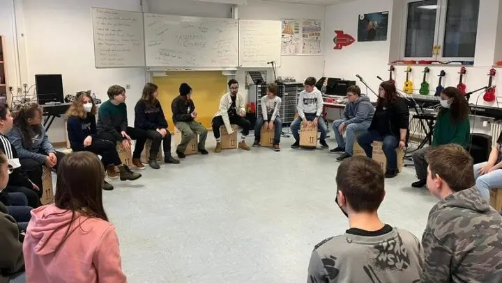 Jugendliche sitzen im Musikraum im Kreis auf Cajóns, an der Tafel steht „Rassismus & Courage“