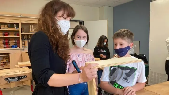 Lehrerin zeigt zwei Kindern mit Masken ein Holzbrett im Werkraum