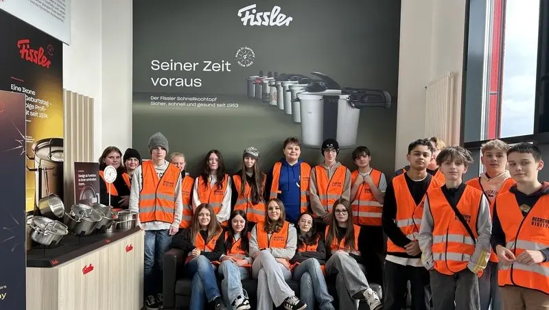 Schülergruppe in orangefarbenen Besucherwesten im Fissler-Showroom vor einer Wand mit Schnellkochtöpfen