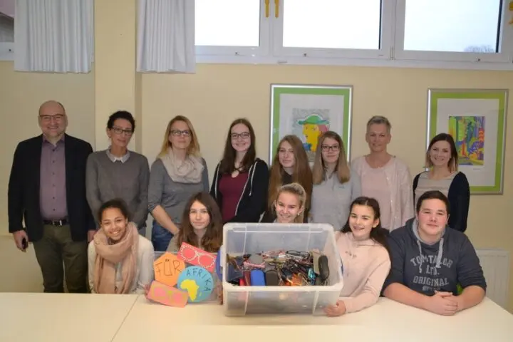 Schülerinnen, Schüler und Erwachsene hinter einer Kiste voller gespendeter Brillen und Schildern „Für Afrika“