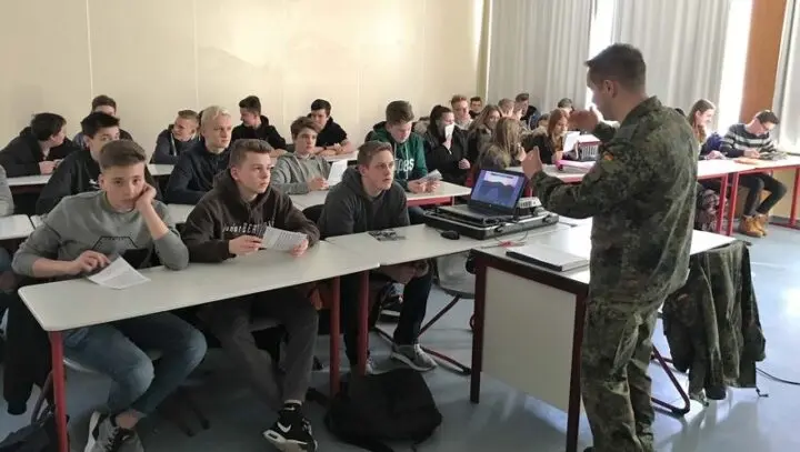 Soldat in Tarnuniform hält vor Schülerinnen und Schülern im Klassenraum einen Vortrag