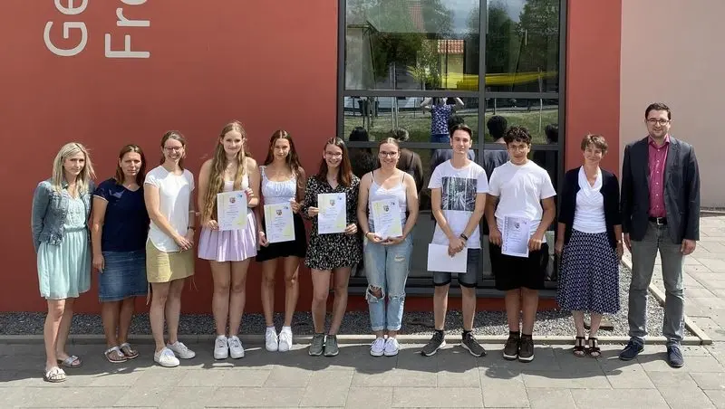 Schülerinnen und Schüler mit Urkunden stehen mit Erwachsenen vor dem Schulgebäude