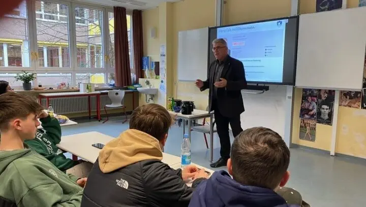 Mann im Anzug spricht vor Jugendlichen im Klassenraum, auf dem Bildschirm „Herzlich Willkommen“