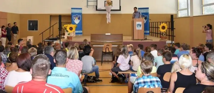 Vollbesetzte Aula bei der Einschulungsfeier, vorne ein Redner am Pult zwischen Sonnenblumen