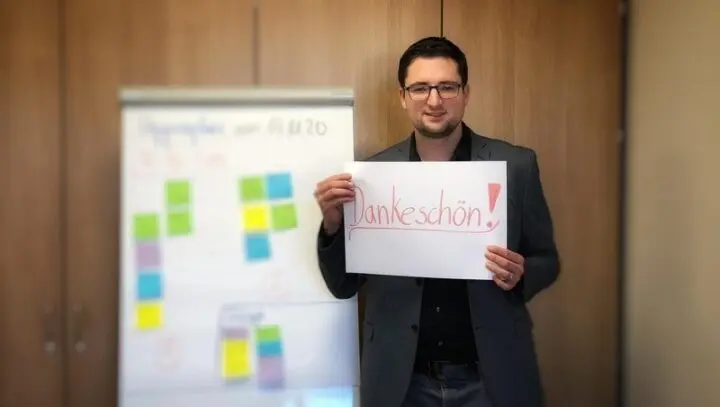 Ein Mann hält ein Blatt mit der Aufschrift „Dankeschön!“ hoch, daneben ein Flipchart