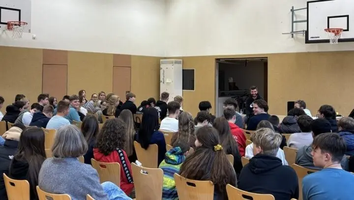 Jugendliche sitzen in Stuhlreihen in der Turnhalle und hören einem Referenten zu