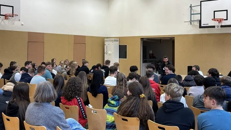 Jugendliche sitzen in Stuhlreihen in der Turnhalle und hören einem Referenten zu