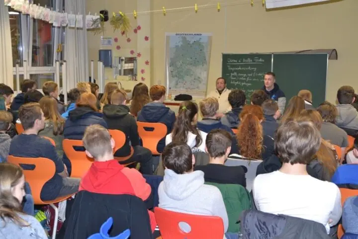 Zwei Männer sitzen vor einer Tafel und sprechen zu vielen Jugendlichen im Klassenraum