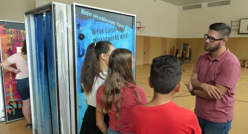 Drei Jugendliche an der Ausstellungsstation „Hinter verschlossenen Türen: Wenn Liebe missbraucht wird!“, ein Mann erklärt