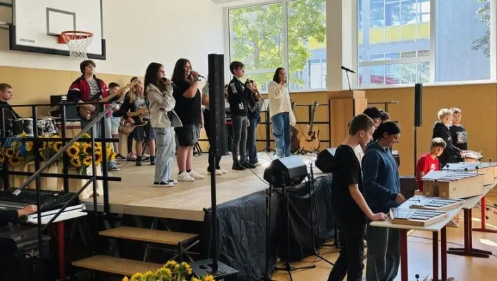 Schülerband mit Gesang und Gitarren auf der Bühne der Turnhalle, davor Jugendliche an Stabspielen