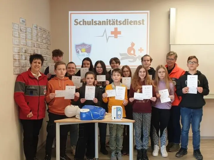 Schulsanitäterinnen und -sanitäter mit Teilnahmebescheinigungen vor dem Schild „Schulsanitätsdienst“, vorne eine Übungspuppe
