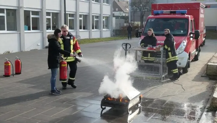 Eine Person löscht unter Anleitung eines Feuerwehrmanns ein Übungsfeuer mit dem Feuerlöscher