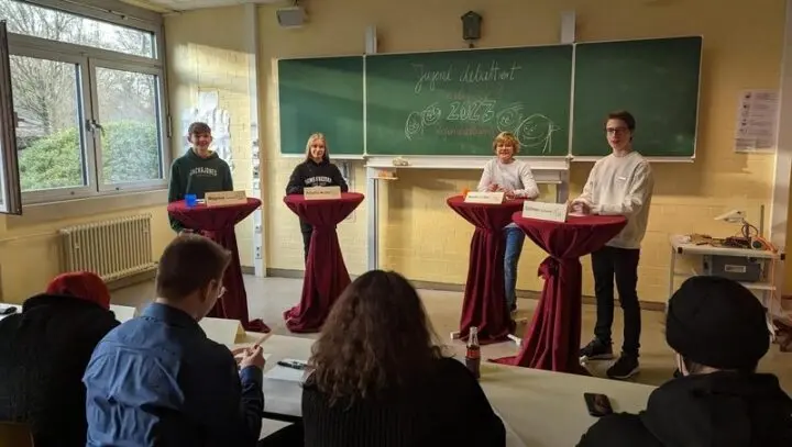 Vier Jugendliche an Stehtischen mit Namensschildern vor einer Tafel „Jugend debattiert 2023“, im Vordergrund Publikum