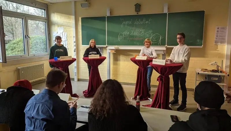 Vier Jugendliche an Stehtischen mit Namensschildern vor einer Tafel „Jugend debattiert 2023“, im Vordergrund Publikum