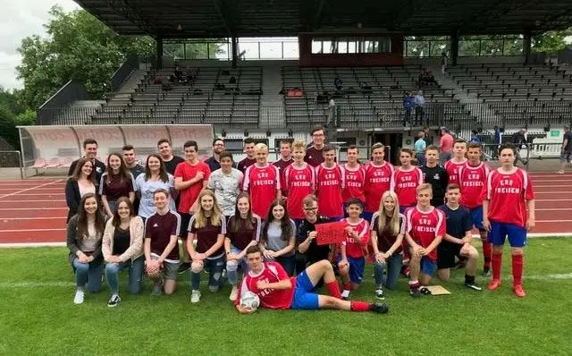 Mannschaftsfoto der Fußballer in roten Trikots mit Fans auf dem Rasen eines Stadions