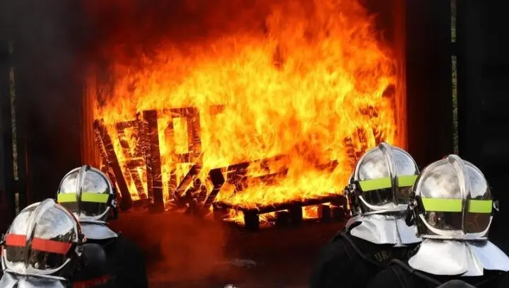 Feuerwehrleute mit silbernen Helmen vor brennenden Holzpaletten
