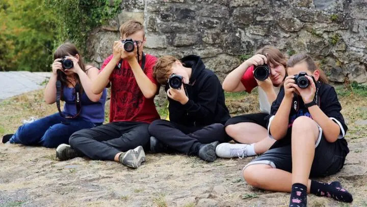 Fünf Jugendliche sitzen auf Steinpflaster vor einer Burgmauer und fotografieren in Richtung Kamera
