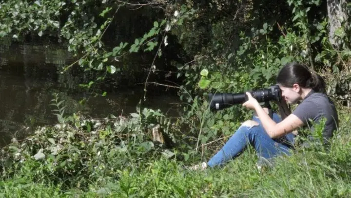Eine Jugendliche sitzt im Grünen am Ufer und fotografiert mit großem Teleobjektiv