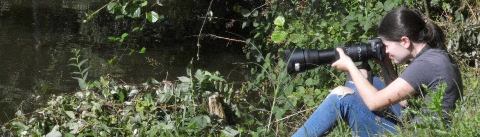 Eine Person sitzt im Grünen am Wasser und fotografiert mit einem großen Teleobjektiv