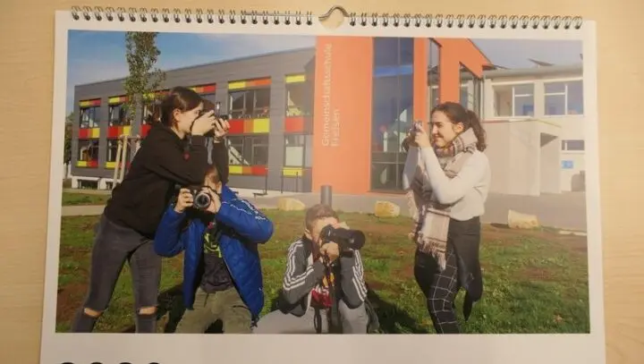Wandkalender, aufgeschlagen mit Foto von vier Jugendlichen mit Kameras vor dem Schulgebäude
