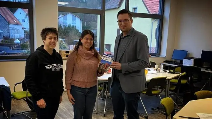 Drei Erwachsene in einem Klassenraum, eine Person hält ein Jahrbuch 2021 in der Hand