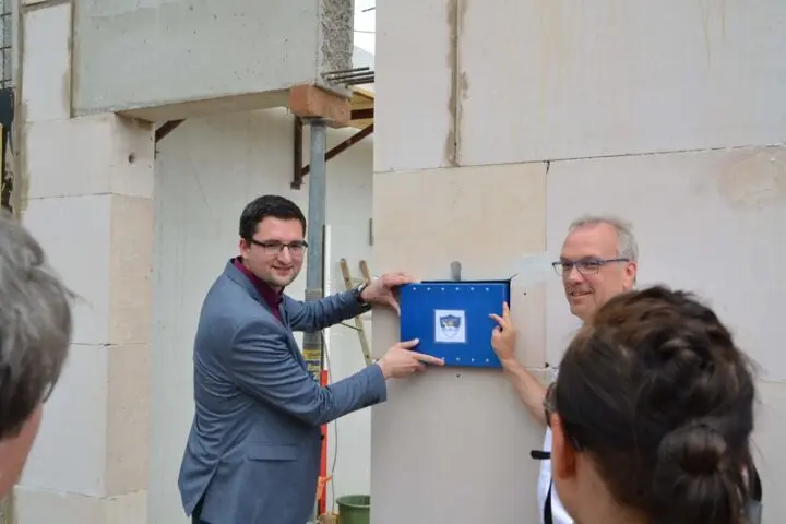 Zwei Männer setzen eine blaue Kassette mit Schullogo in eine Wand aus Mauersteinen ein