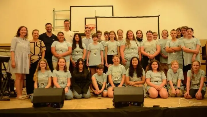 Gruppenfoto der Klasse in einheitlichen grauen T-Shirts mit Erwachsenen auf der Bühne der Turnhalle