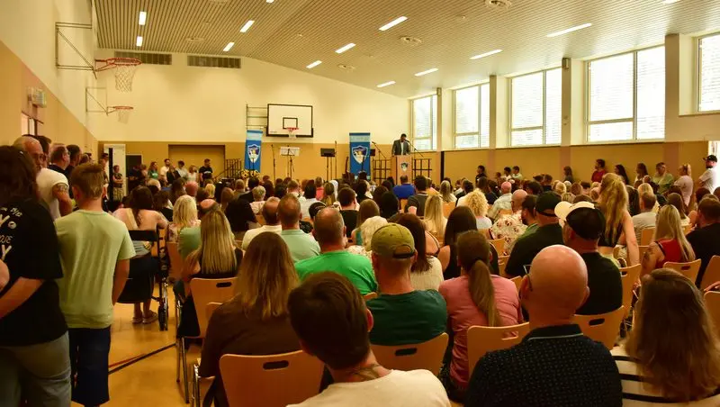 Vollbesetzte Turnhalle bei der Aufnahmefeier, vorne ein Redner am Pult zwischen zwei Schulbannern