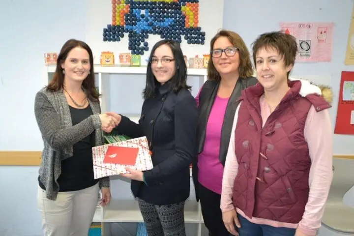 Eine Schülerin erhält mit Handschlag ein Geschenk, daneben drei Frauen in einem Klassenraum