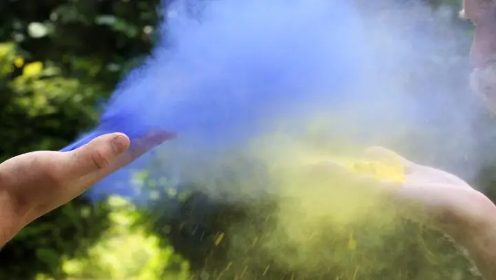 Blaues und gelbes Farbpulver wird von zwei Händen in die Luft gepustet