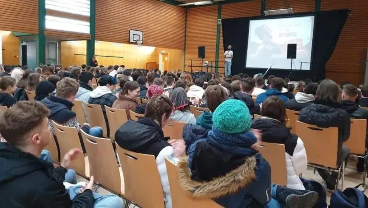 Vollbesetzte Turnhalle, Schülerinnen und Schüler verfolgen einen Vortrag vor einer Leinwand