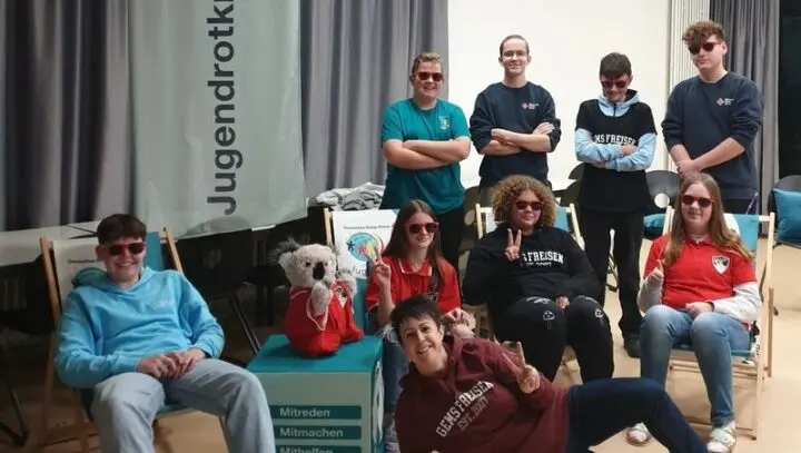 Gruppe Jugendlicher mit roten Sonnenbrillen vor einem Jugendrotkreuz-Banner, ein Würfel mit „Mitreden, Mitmachen“