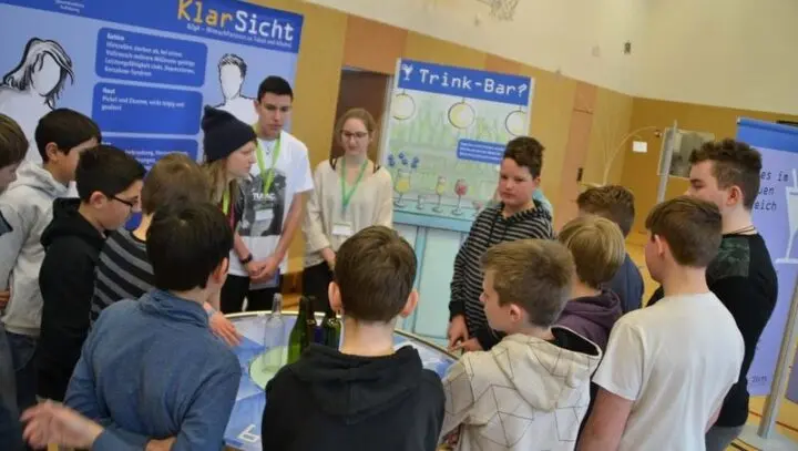 Schülergruppe an der Station „Trink-Bar?“ des KlarSicht-Parcours in der Turnhalle, Betreuende mit Namensschildern