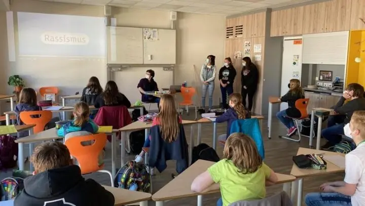 Klasse mit Masken im Klassenraum, drei Schülerinnen stehen vorne, an der Wand die Projektion „Rassismus“