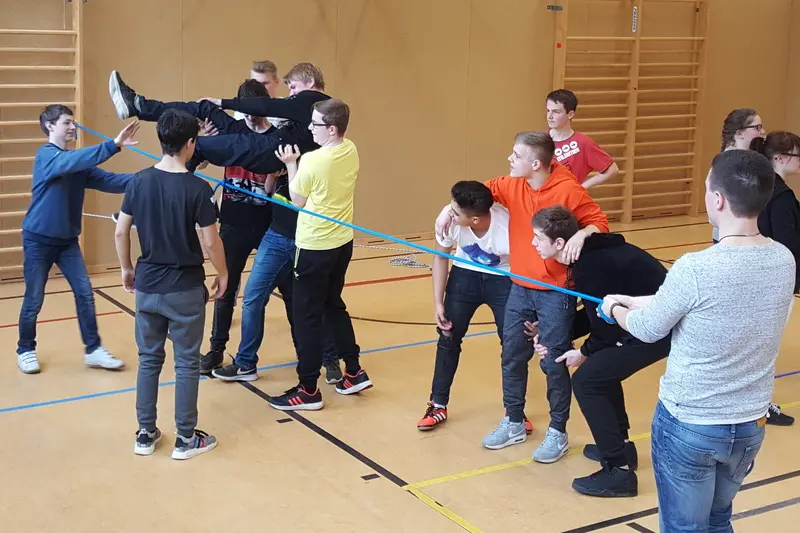 Jugendliche in der Turnhalle bei einem Kooperationsspiel, eine Person wird über ein gespanntes Seil gehoben