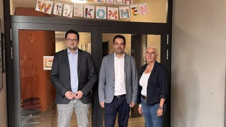 Drei Erwachsene vor einer Glastür mit bunten Buchstaben „Willkommen“
