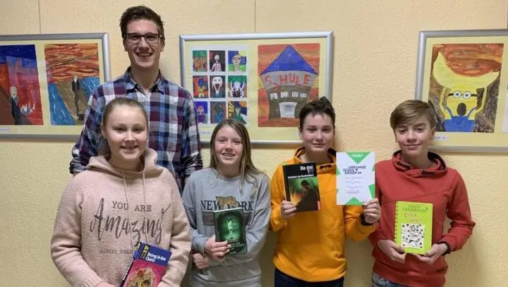 Vier Schülerinnen und Schüler mit Büchern und einer Urkunde, dahinter ein junger Mann, an der Wand Kinderbilder