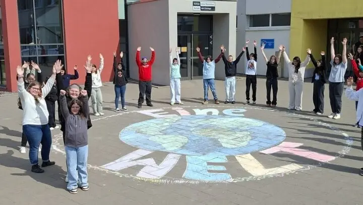 Schülerinnen und Schüler stehen mit erhobenen Armen um ein Kreidebild der Erde mit „Eine Welt“ auf dem Schulhof