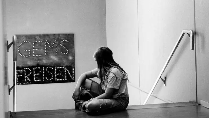 Schwarz-Weiß-Foto: Schülerin sitzt im Treppenhaus auf dem Boden vor einem Leuchtschild „GEMS FREISEN“