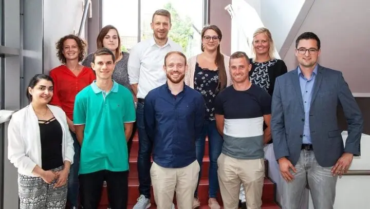 Neun Personen stehen in zwei Reihen auf einer Treppe für ein Gruppenfoto