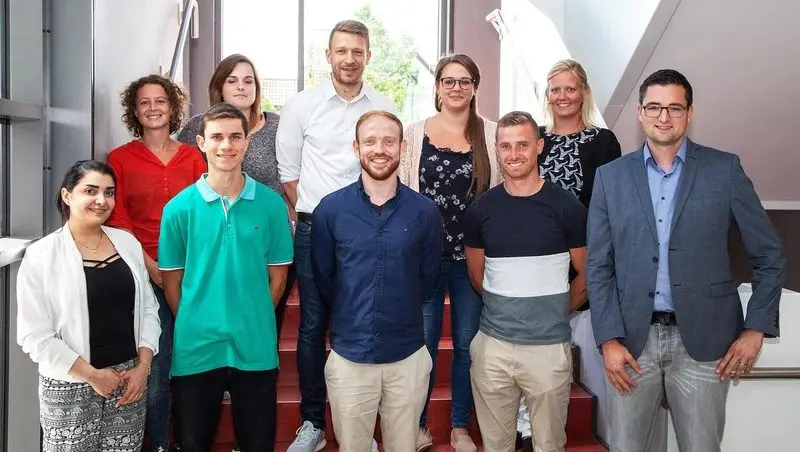 Neun Personen stehen in zwei Reihen auf einer Treppe für ein Gruppenfoto