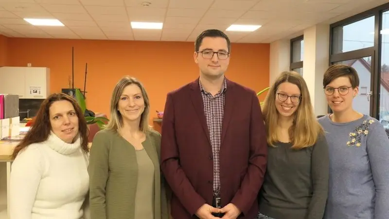 Fünf Lehrkräfte stehen nebeneinander in einem Raum mit orangefarbener Wand